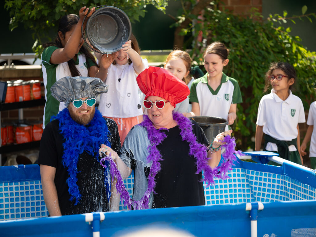 Two teachers wearing shower caps in padding pool while pupils tip milk over them